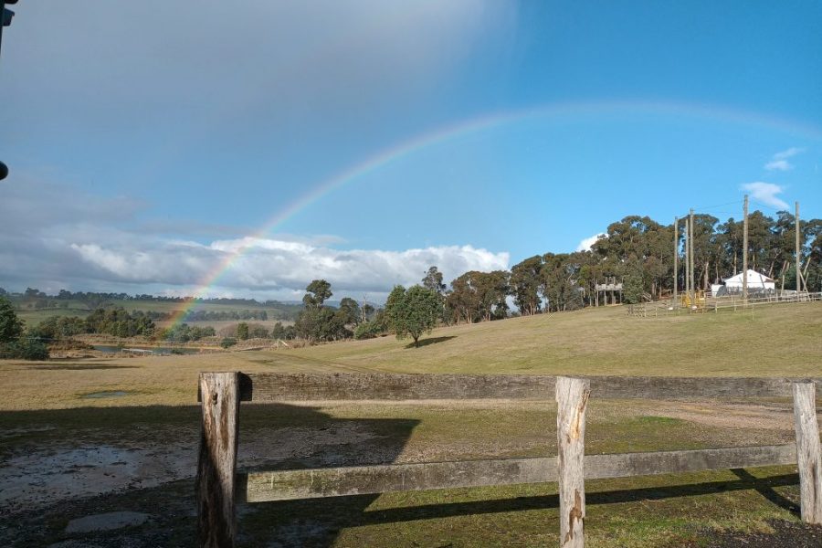 A rainbow arcs over a grassy field with scattered trees, viewed from behind a wooden fence under a partly cloudy sky at a PGL camp.