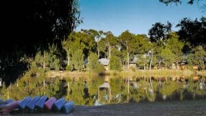 Calm lake reflecting trees with stacked canoes on the shore under clear sky.