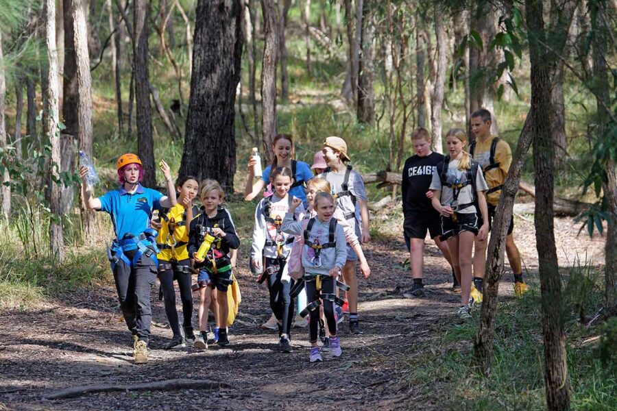 A group of children and two adults hiking on a forest trail, one adult leading and pointing, some children equipped with helmets.