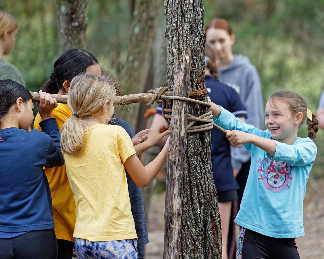 Group of children engaging in a team-building activity, tying ropes around a tree in a forest setting.