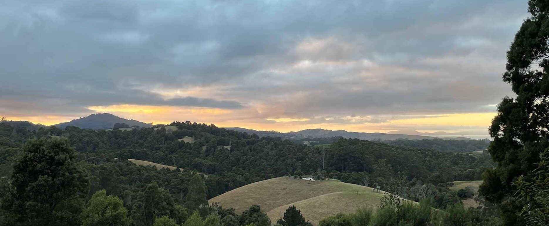 A scenic view of rolling hills at sunset, featuring a mix of trees and open fields under a partly cloudy sky.