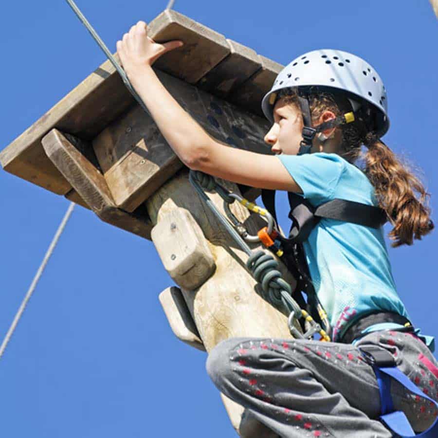 A young girl in a helmet and harness climbs a wooden obstacle at a group camp against a clear blue sky.