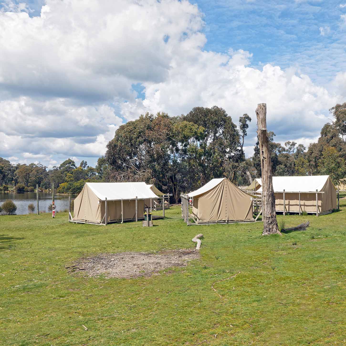 Canvas tents set up on a grassy field near a lake with trees and a partly cloudy sky in the background.