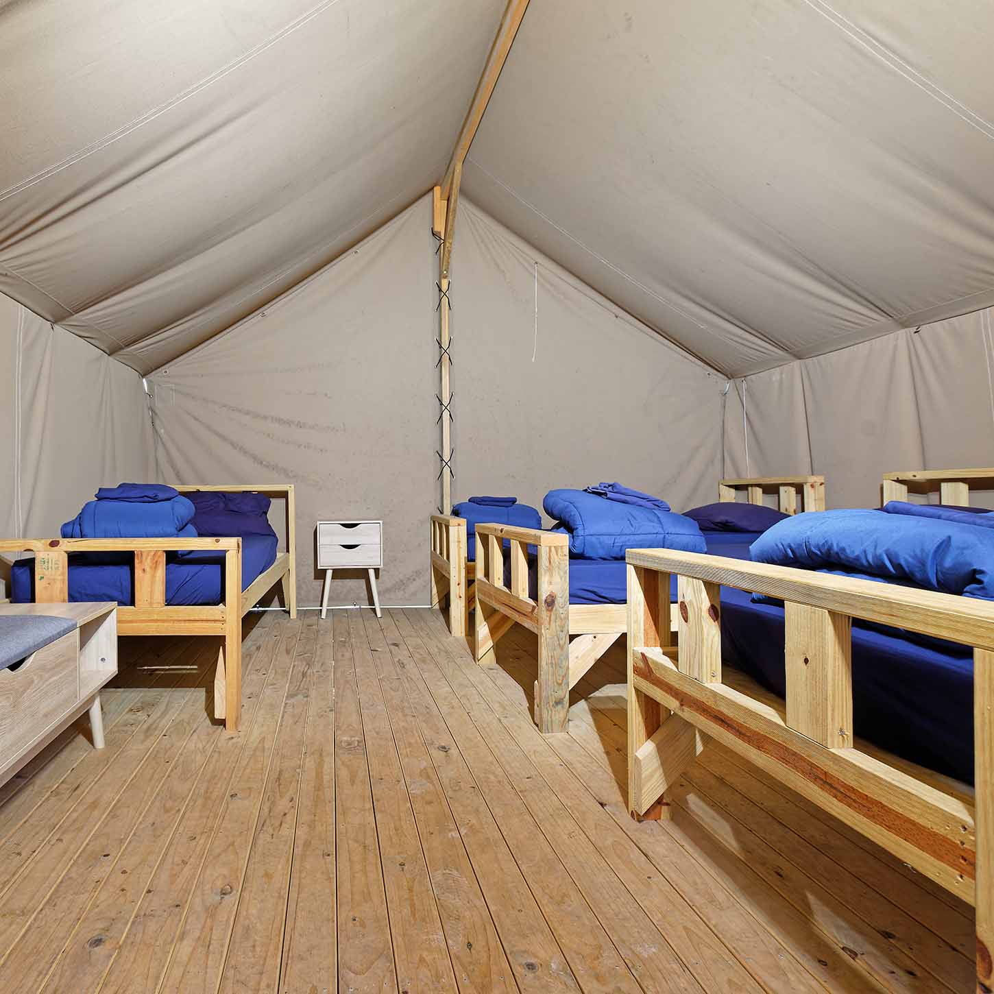 Interior of a large tent with multiple wooden bunk beds covered in blue bedding, a small white bedside table on a wooden floor.