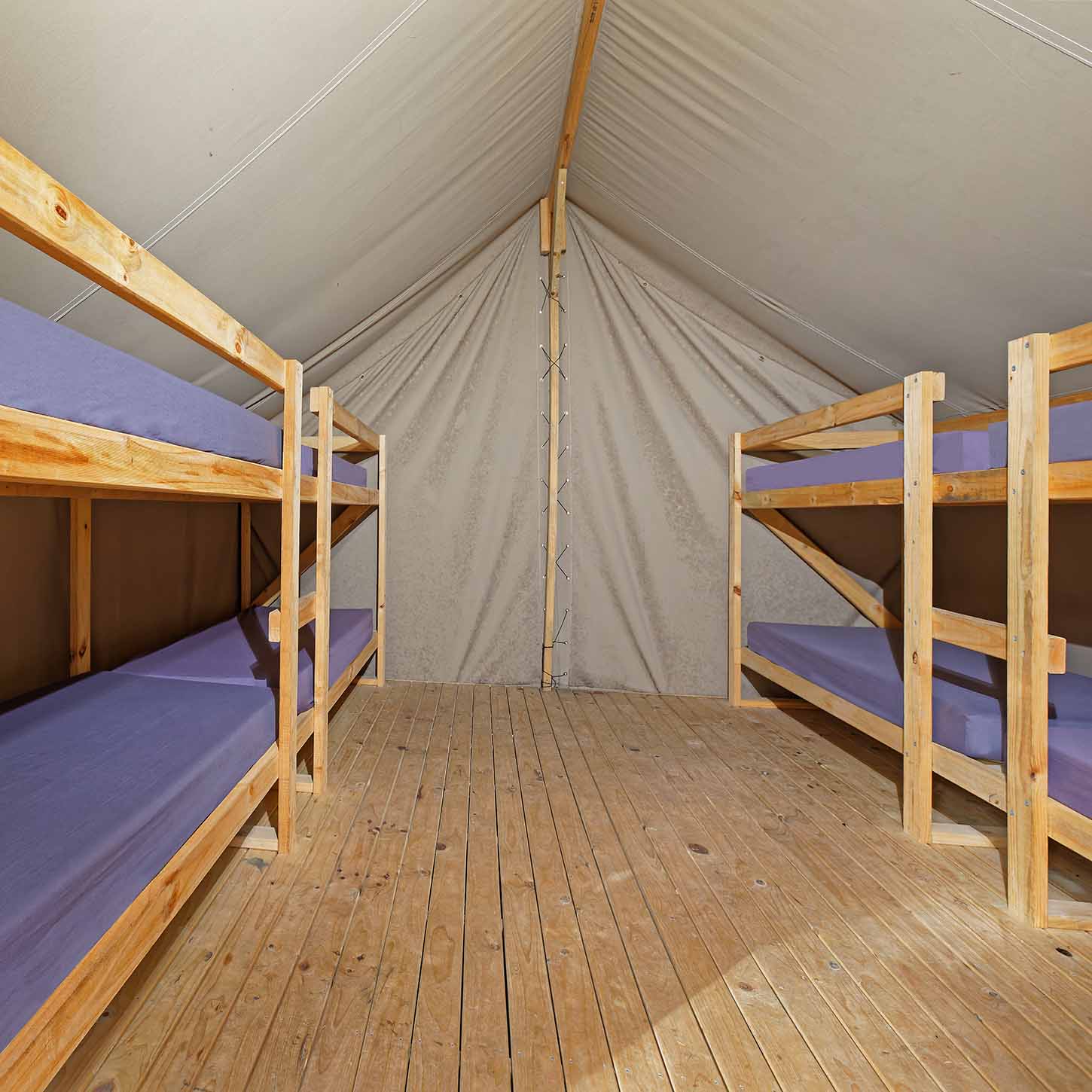 Interior of a canvas tent with four wooden bunk beds and a wooden floor, illuminated by natural light.