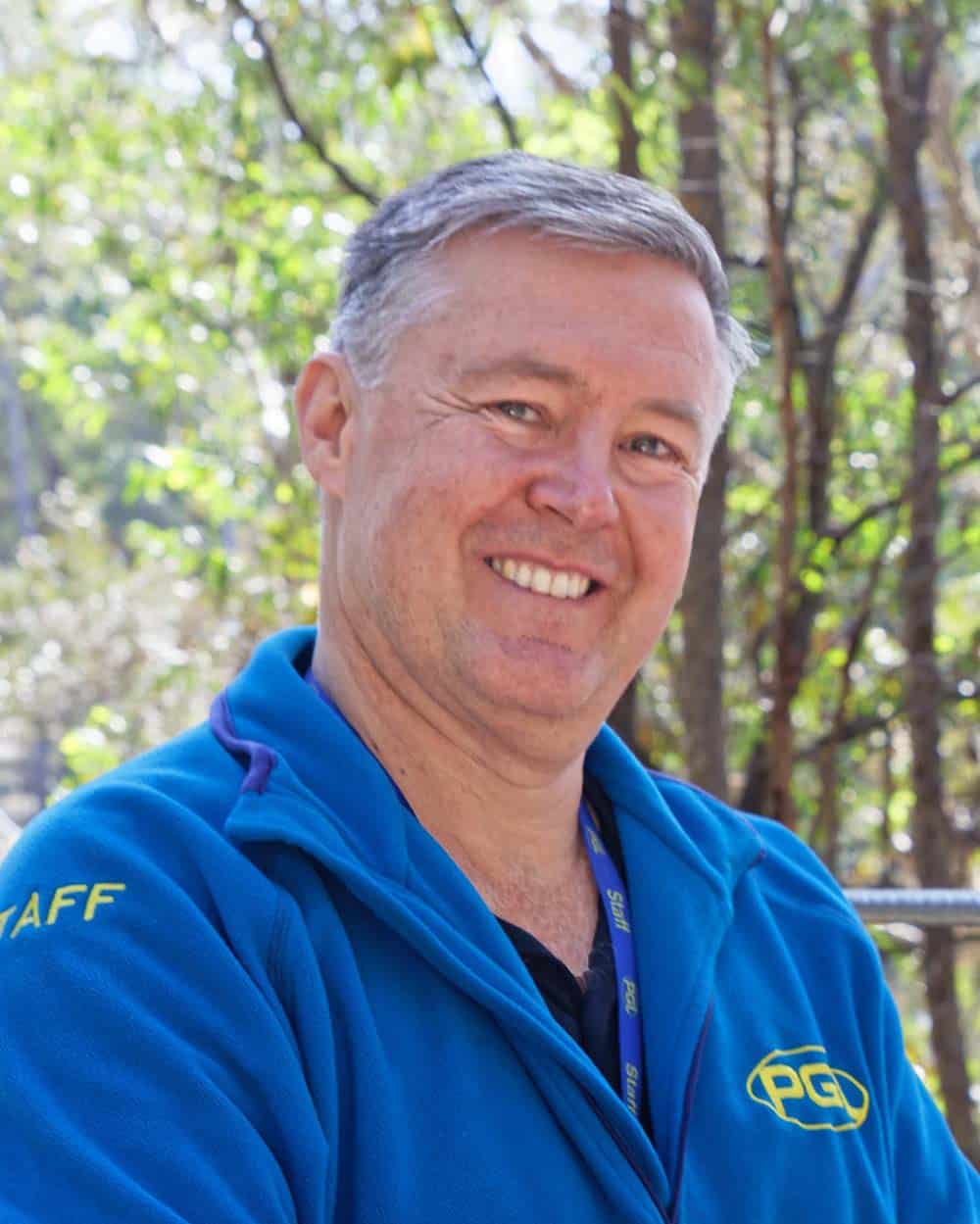 Middle-aged man wearing a blue staff hoodie and a lanyard, smiling in a wooded outdoor setting.