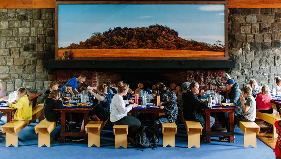 People dining at long tables inside a spacious room with a large landscape painting on the wall.