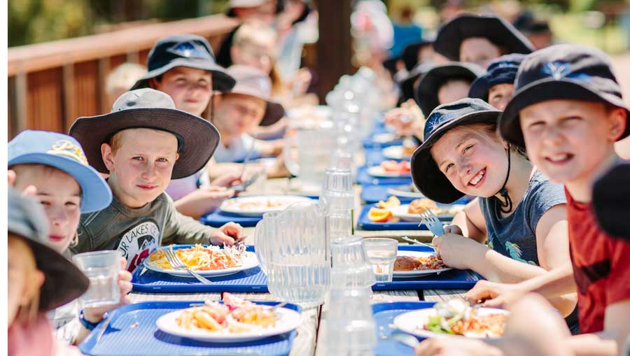 Group of children wearing sun hats eating lunch at a long outdoor table on a sunny day.