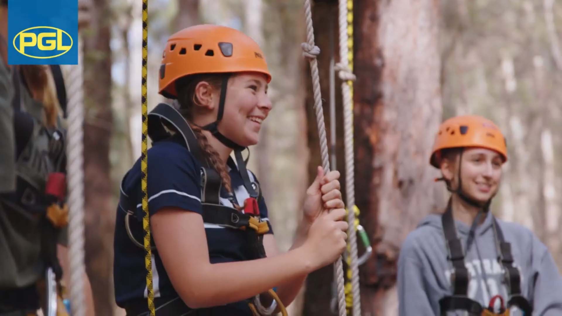 Teen girls in safety gear preparing for a ropes course in a forest setting, smiling and holding onto ropes.