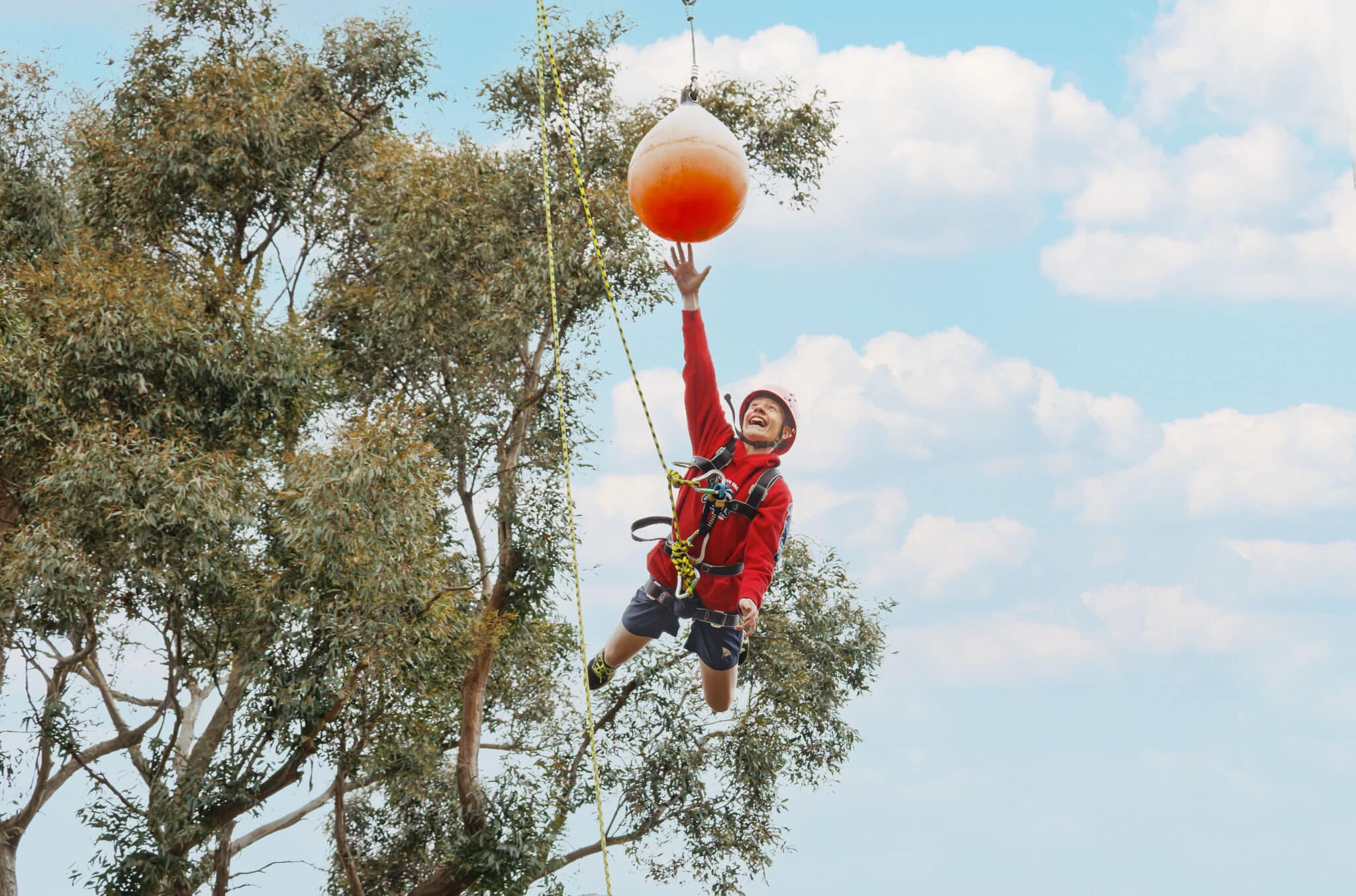 A child in a harness at PGL camps joyfully reaches for a hanging buoyant ball against a backdrop of blue sky and trees.