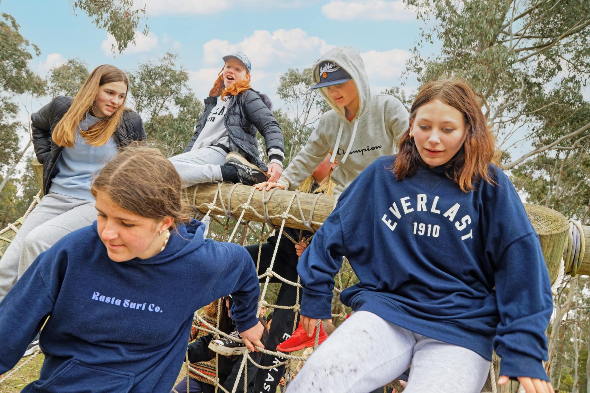 Group of teenagers sitting and climbing on a fallen tree at an Easter Holiday Camp, interacting casually in a natural outdoor setting.