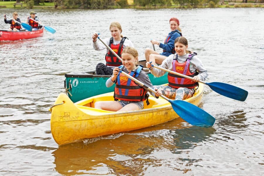 Group of young people wearing life jackets paddling canoes at a PGL camp on a river, smiling and looking at the camera.