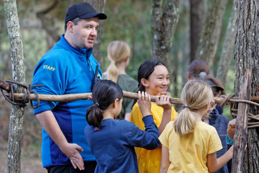 A staff member in a blue shirt and cap teaches a group of diverse children how to build a structure with sticks in a forest setting.