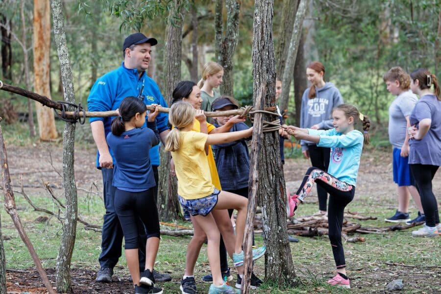 Children and an adult superviser building a structure with sticks between trees in a wooded area.