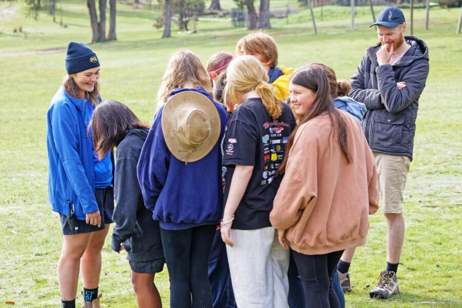A group of young people in casual attire and varying headgear stand in a grassy field, engaged in conversation with smiles.