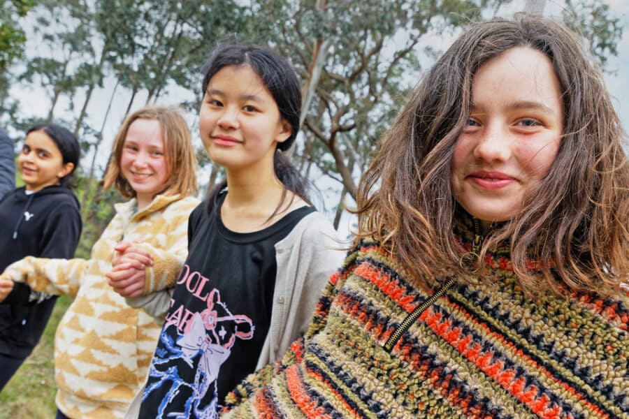 A group of diverse teenage girls smiling and standing outdoors, with one girl in a colorful sweater looking at the camera.