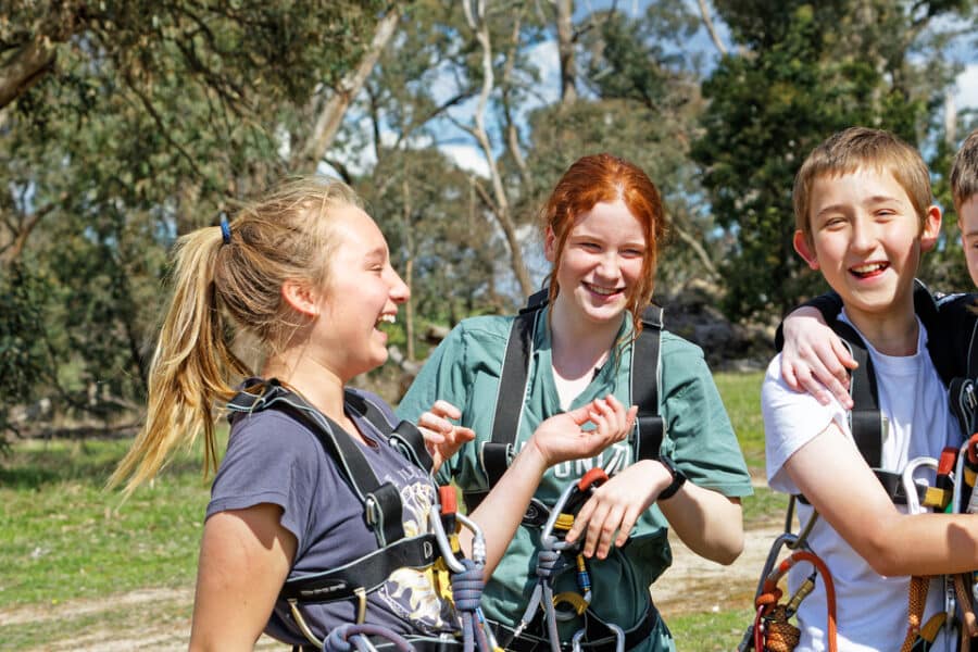 Four smiling children outdoors, wearing harnesses and holding ropes, stand together under trees in a sunny park.