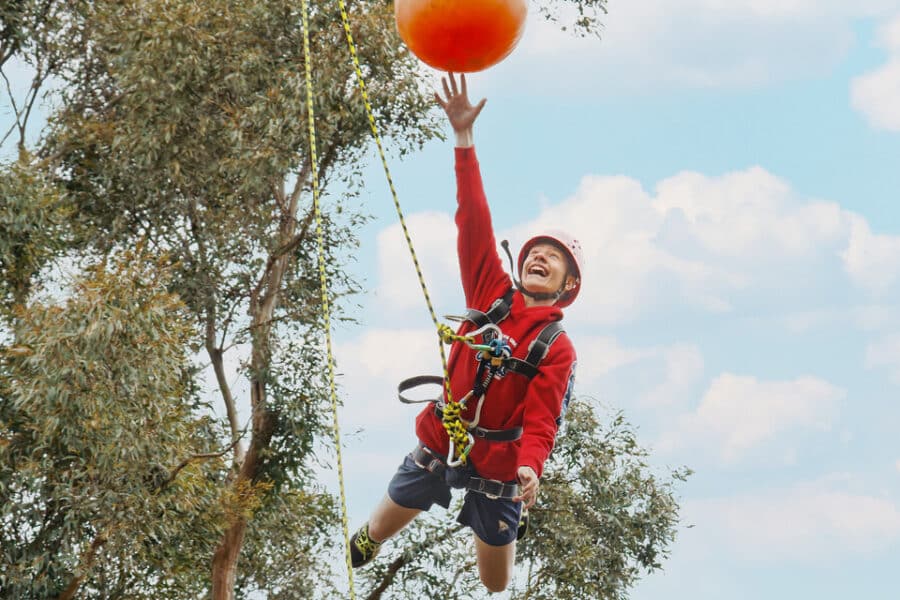 A young boy in a harness and helmet joyously reaching for a large orange ball while zip-lining against a blue sky and trees.