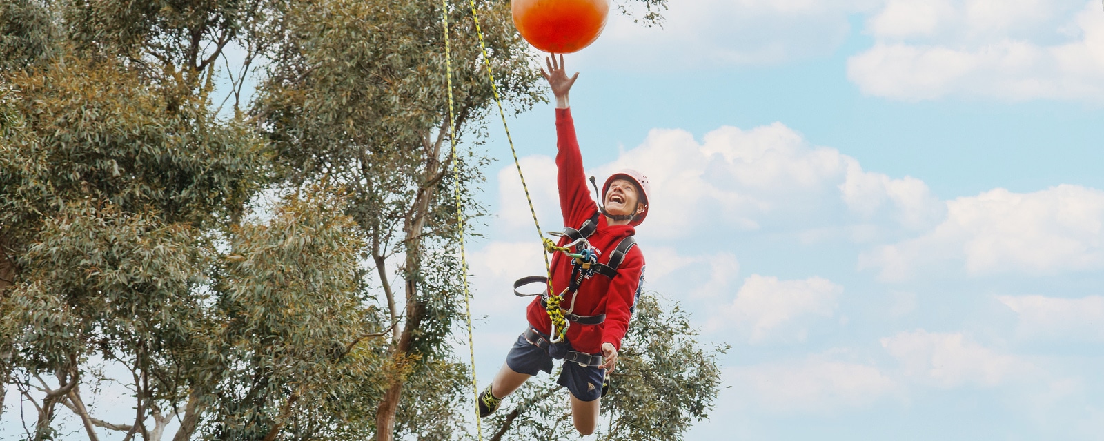 A young boy in a harness and helmet joyously reaching for a large orange ball while zip-lining against a blue sky and trees.