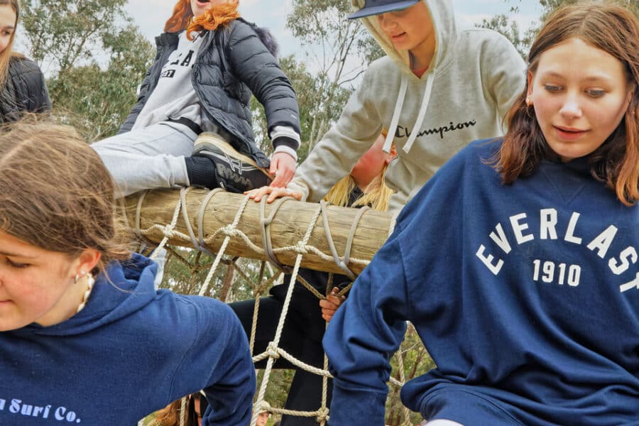 Group of young people in casual wear climbing and sitting on a rope structure in a park, some focused on adjusting the ropes.