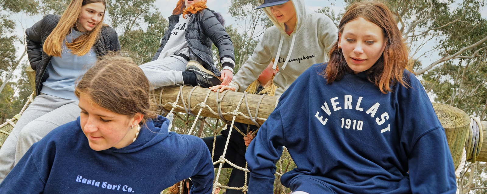 Group of young people in casual wear climbing and sitting on a rope structure in a park, some focused on adjusting the ropes.