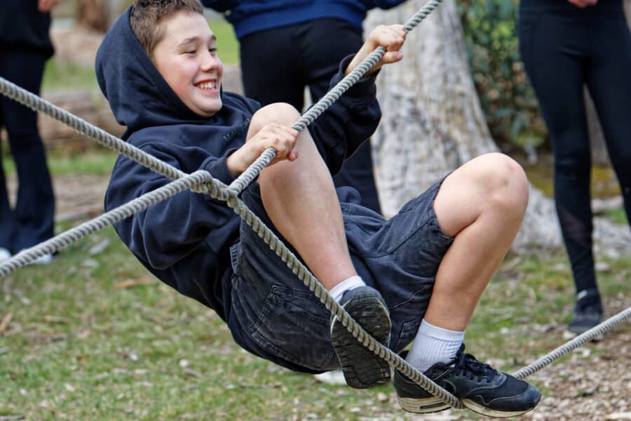 A boy smiles while swinging on a rope at a park, with other children and adults standing in the background.