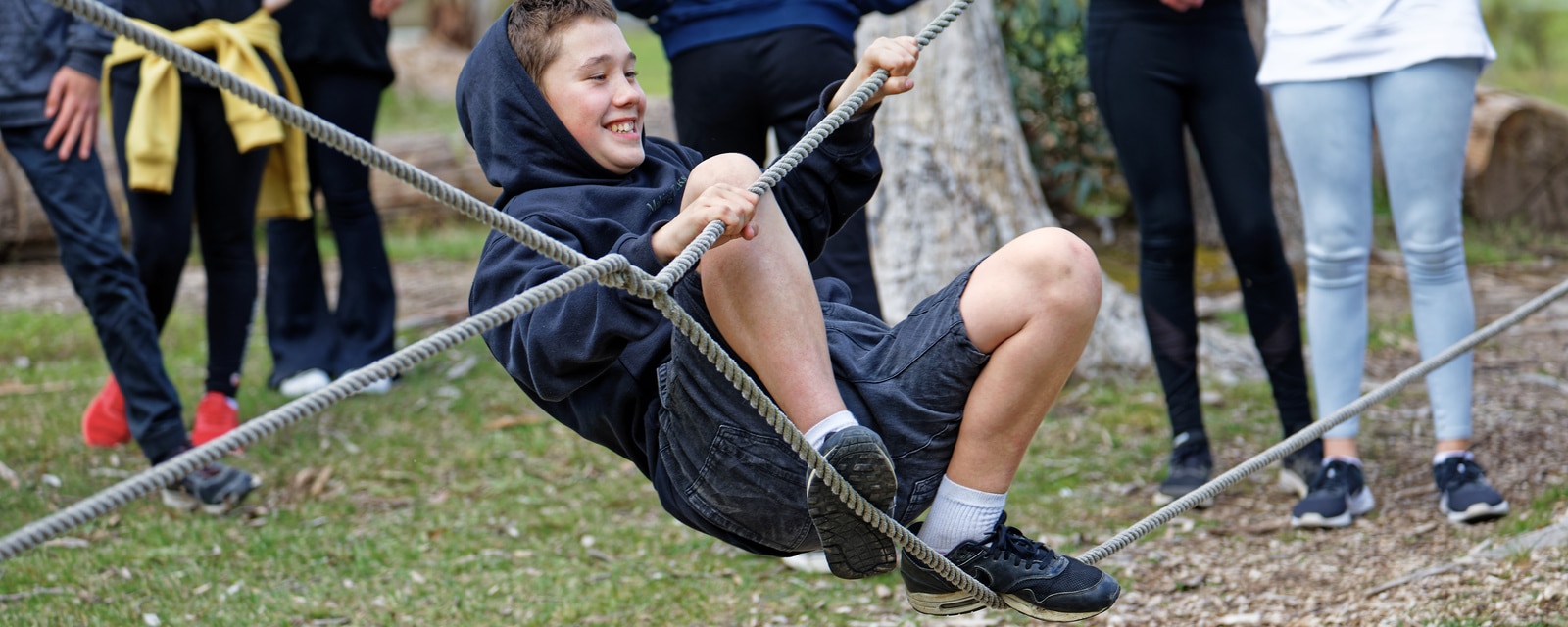 A boy smiles while swinging on a rope at a park, with other children and adults standing in the background.