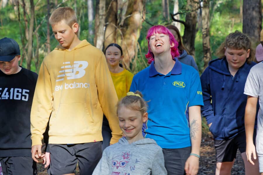 A group of diverse children walking in a forested area, some smiling and talking, others look ahead.