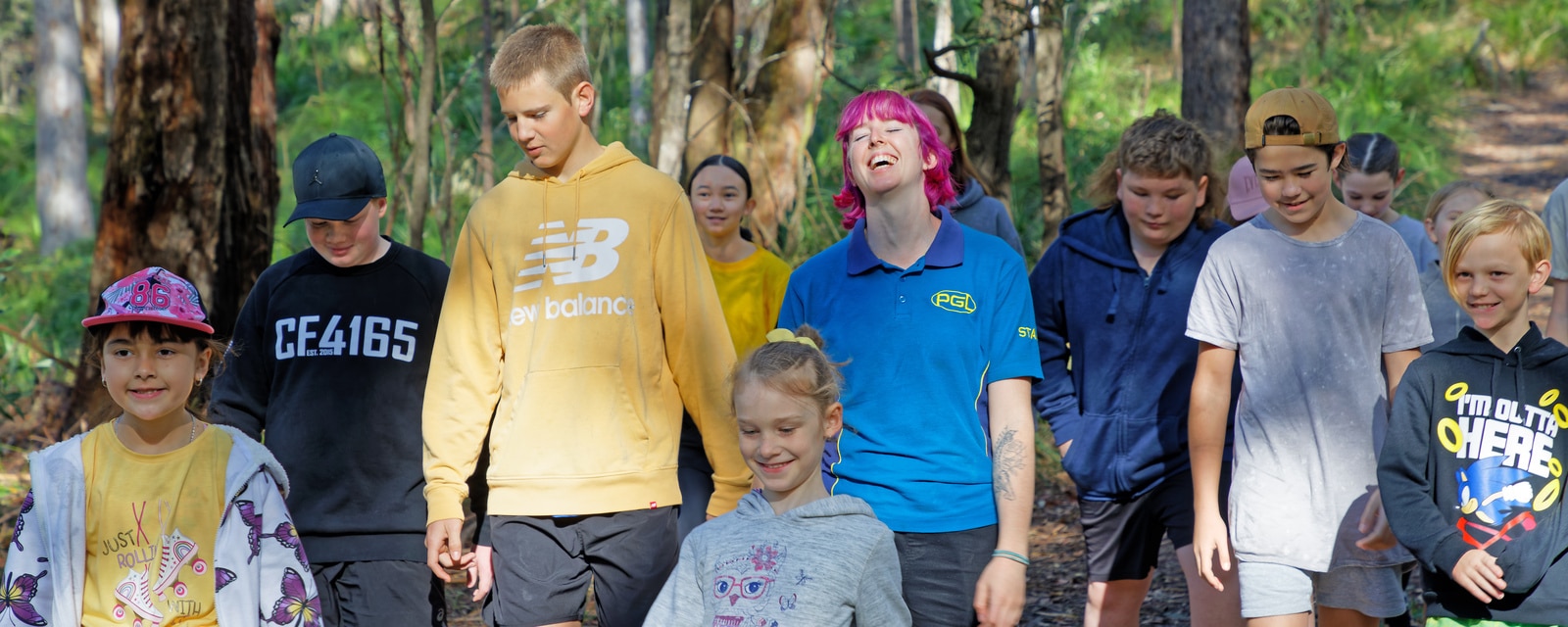 A group of diverse children walking in a forested area, some smiling and talking, others look ahead.