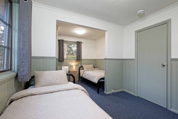 A tidy room featuring two single beds with beige bedding, gray and white walls, a bedside table with a lamp, and a carpeted floor.