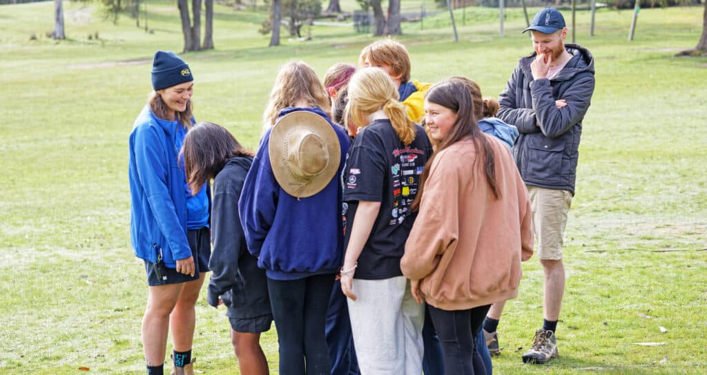 A group of young people and an adult standing on grass, engaging in a conversation, with an instructor wearing scout uniforms and outdoor attire.