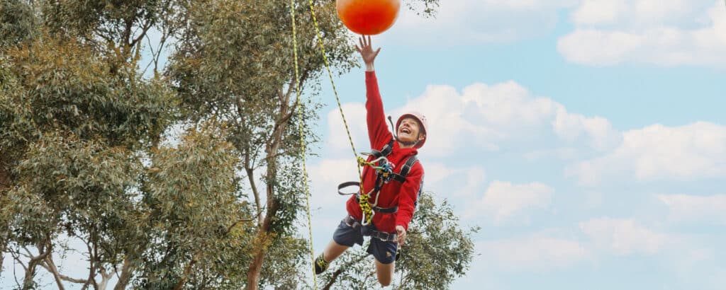 A boy in a red jacket and helmet reaches for a large orange ball while suspended in the air, with trees and a cloudy sky in the background.
