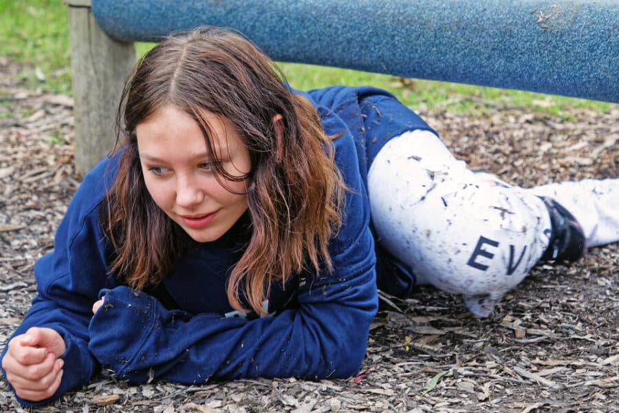 Young girl in a blue sweatshirt and patterned leggings crawling under a low bar at a park, looking focused.