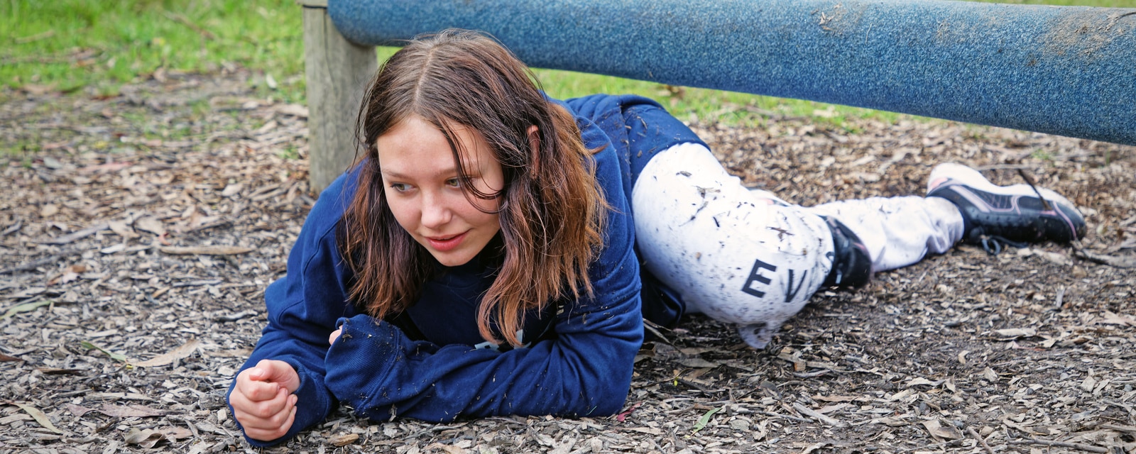 Young girl in a blue sweatshirt and patterned leggings crawling under a low bar at a park, looking focused.