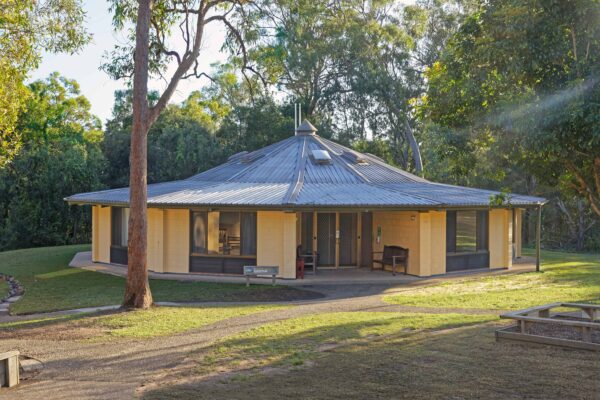 A single-story octagonal building with a metal roof, surrounded by trees and lit by evening sunlight.