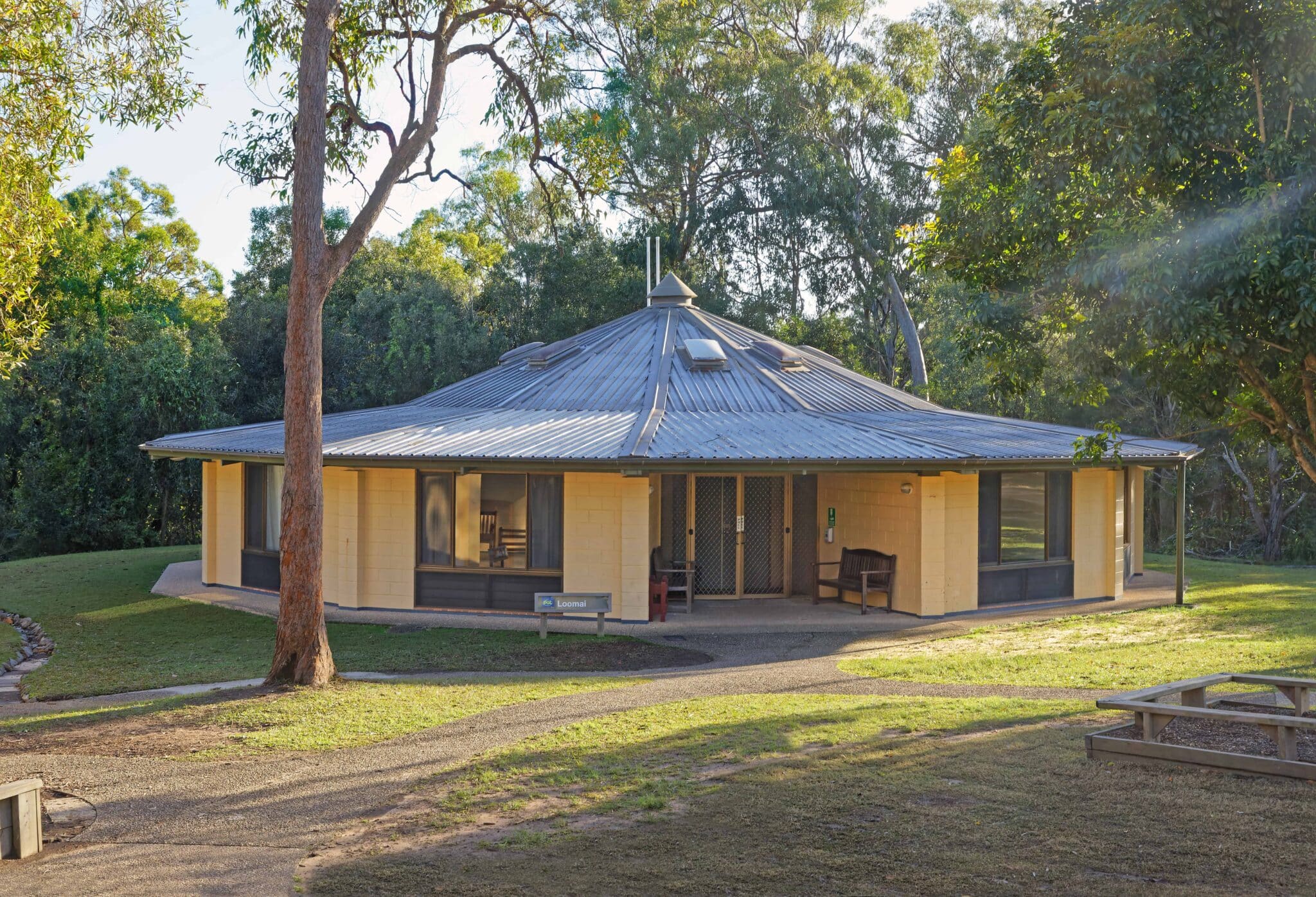 A single-story octagonal building with a metal roof, surrounded by trees and lit by evening sunlight.