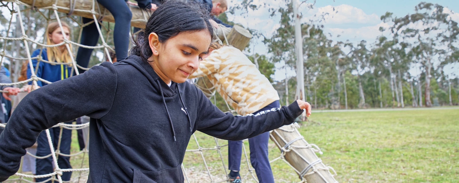 A young girl climbing on a rope structure at a park, with others around and trees in the background.