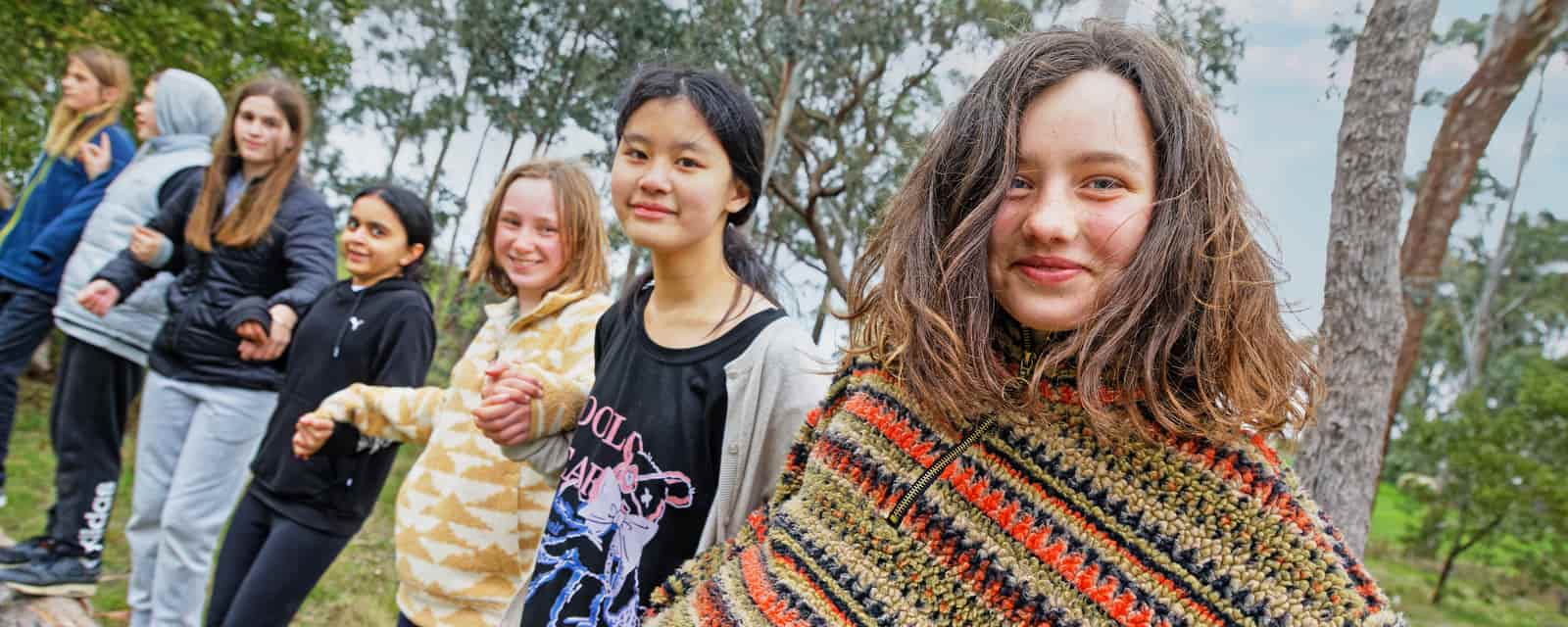 A group of diverse teenagers smiling outdoors, with one girl in a colorful sweater standing prominently in front.