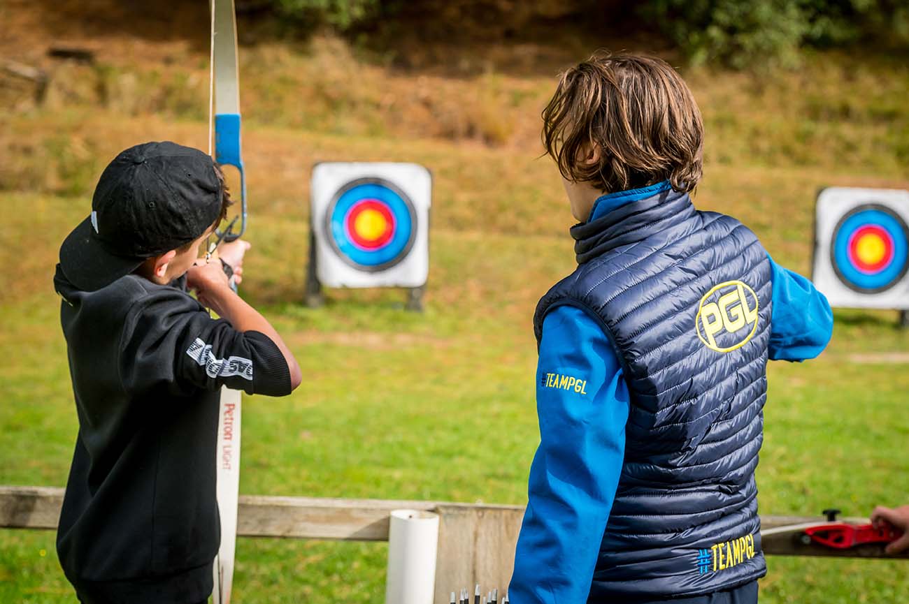 Two boys discover the sport of archery; one is aiming at a target while the other, wearing a blue and black jacket, watches.