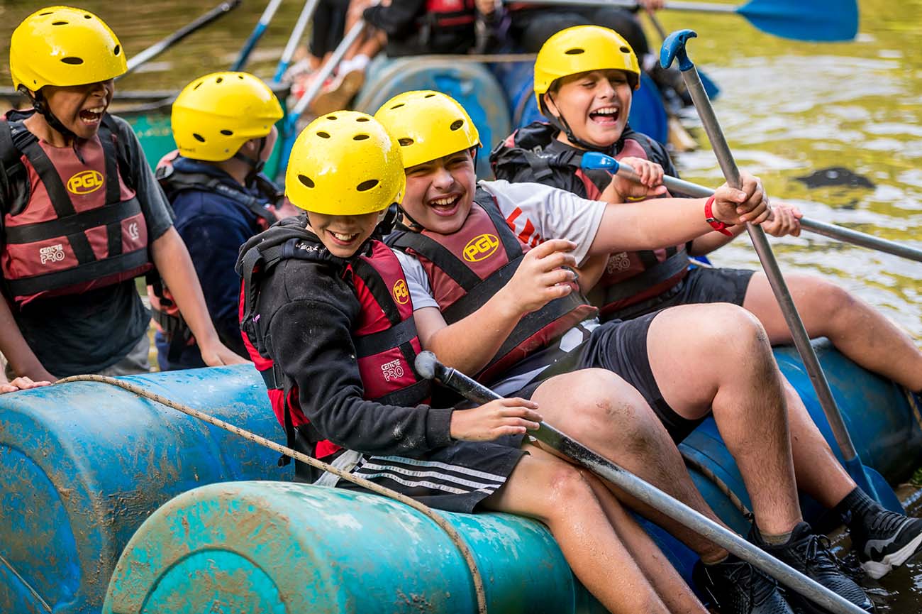 Group of teenagers wearing helmets and life vests laughing and uniting in effort while rowing in a blue raft on a river.