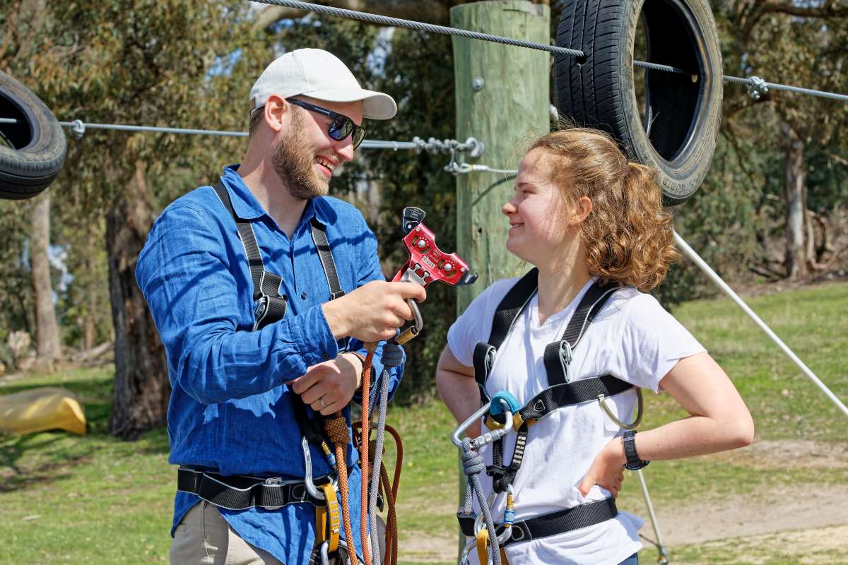 A man in a cap unites climbing gear demonstrations with practical advice for a young girl in a harness at an outdoor adventure park with tire obstacles in the background.