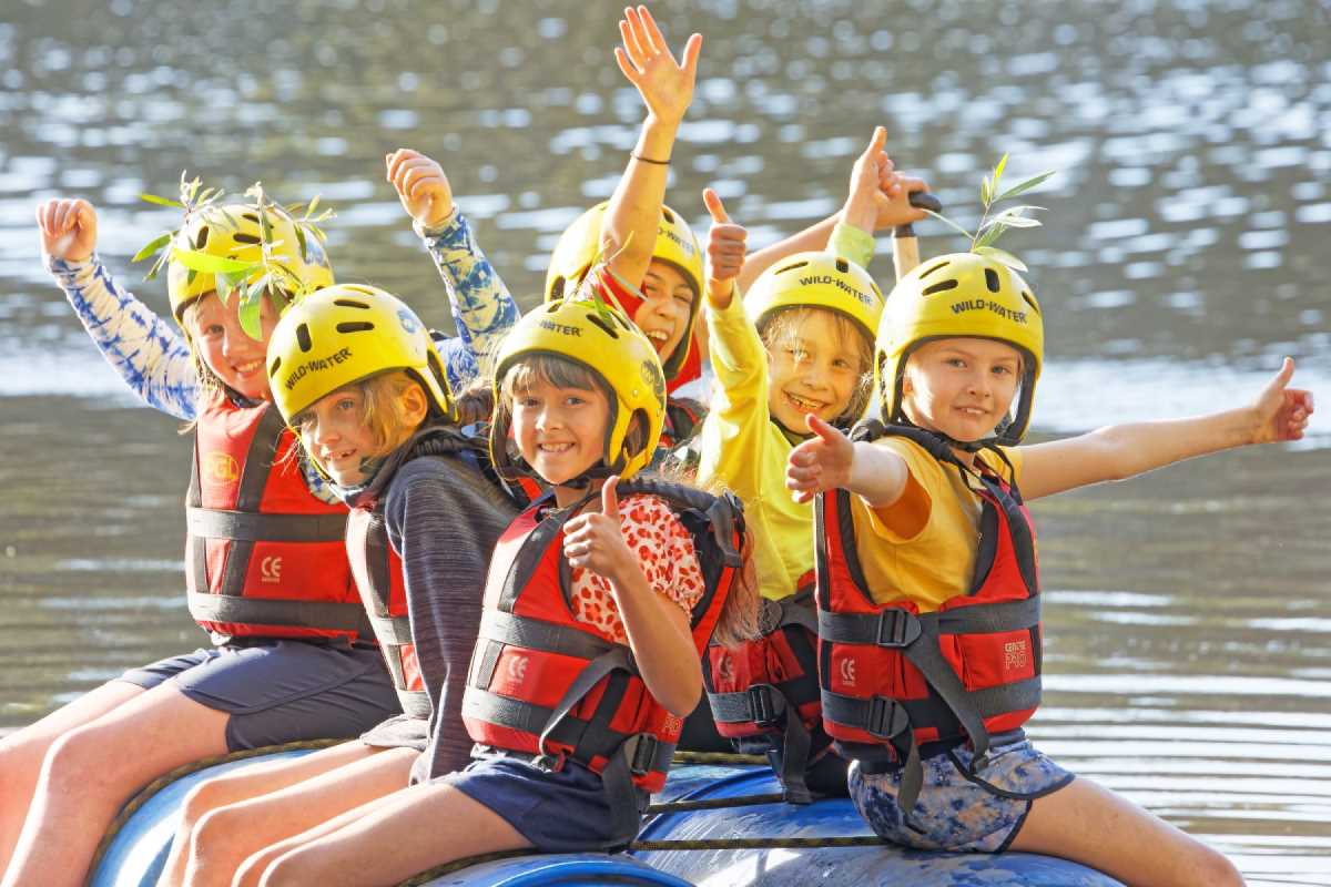 Six children in safety vests and helmets excitedly discover rafting as they pose with thumbs up on a sunny day.