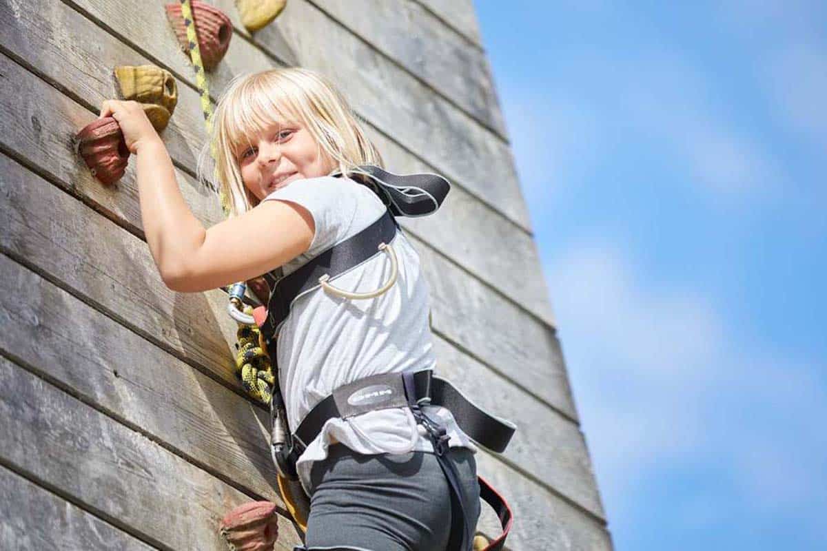 Young girl discovering the joy of climbing an outdoor wall, smiling at the camera, equipped with a harness and rope.