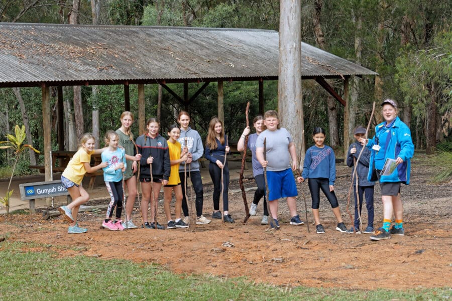 A group of children with a leader wearing a blue jacket, standing cheerfully on a dirt path near a wooden shelter in a park.