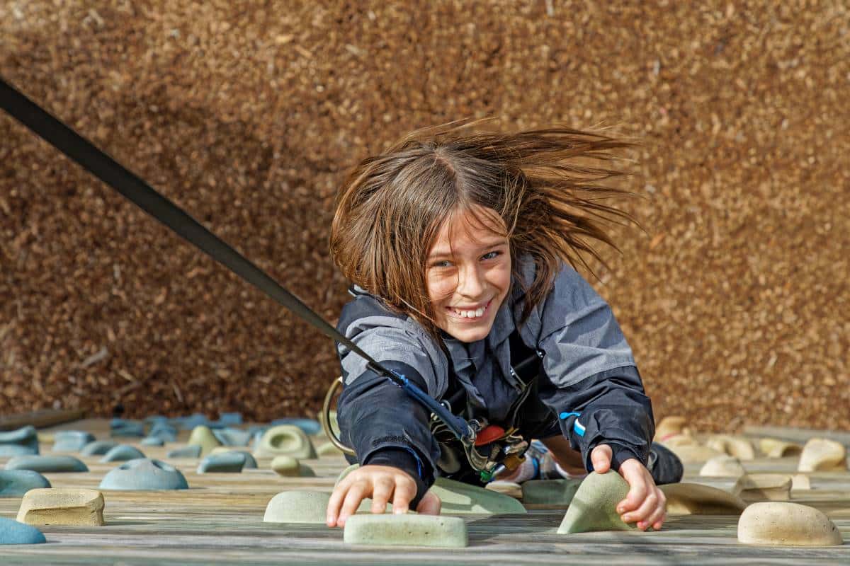 A joyful child with long, flowing hair unites determination and joy as she climbs a rock wall, smiling broadly in a playground setting.