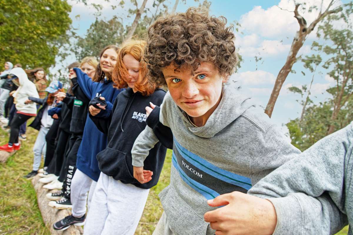 Young person with curly hair playfully pointing at the camera, united with laughing friends in a park.
