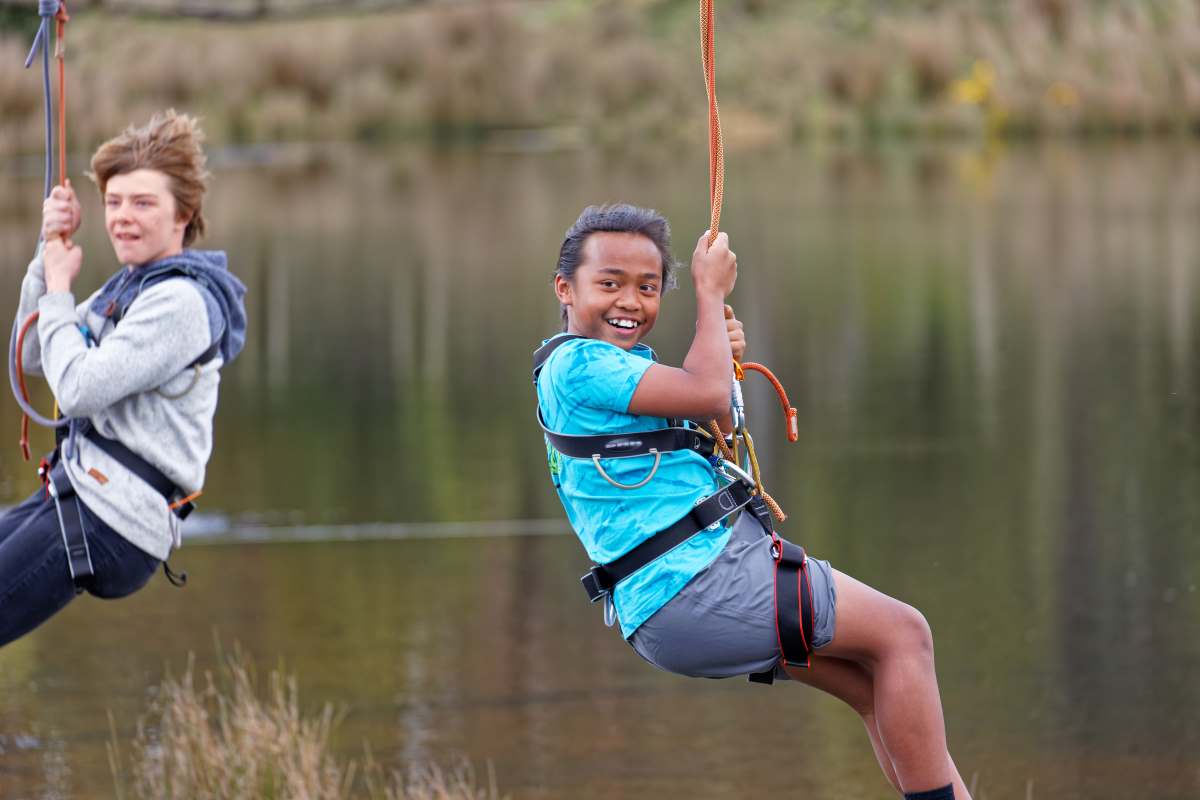 Two children uniting in joy on a zip line over a lake, the girl in front smiling at the camera while the boy trails behind.
