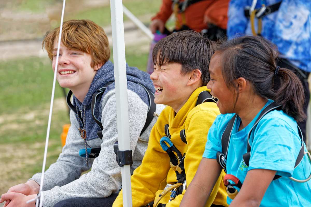 Three kids united in laughter on outdoor swings, two boys and a girl, wearing casual clothes and safety harnesses.