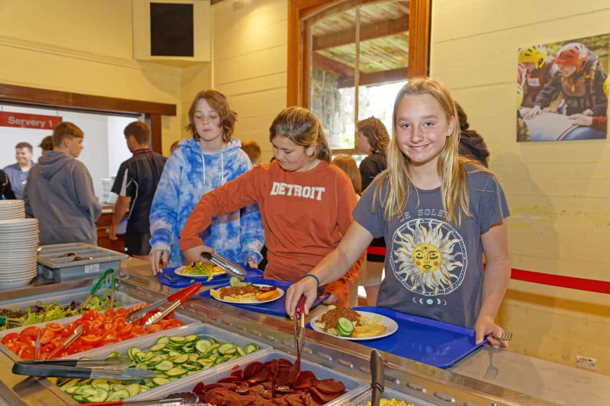 Young students unite as they serve themselves at a school cafeteria salad bar, featuring a selection of fresh vegetables and deli meats.