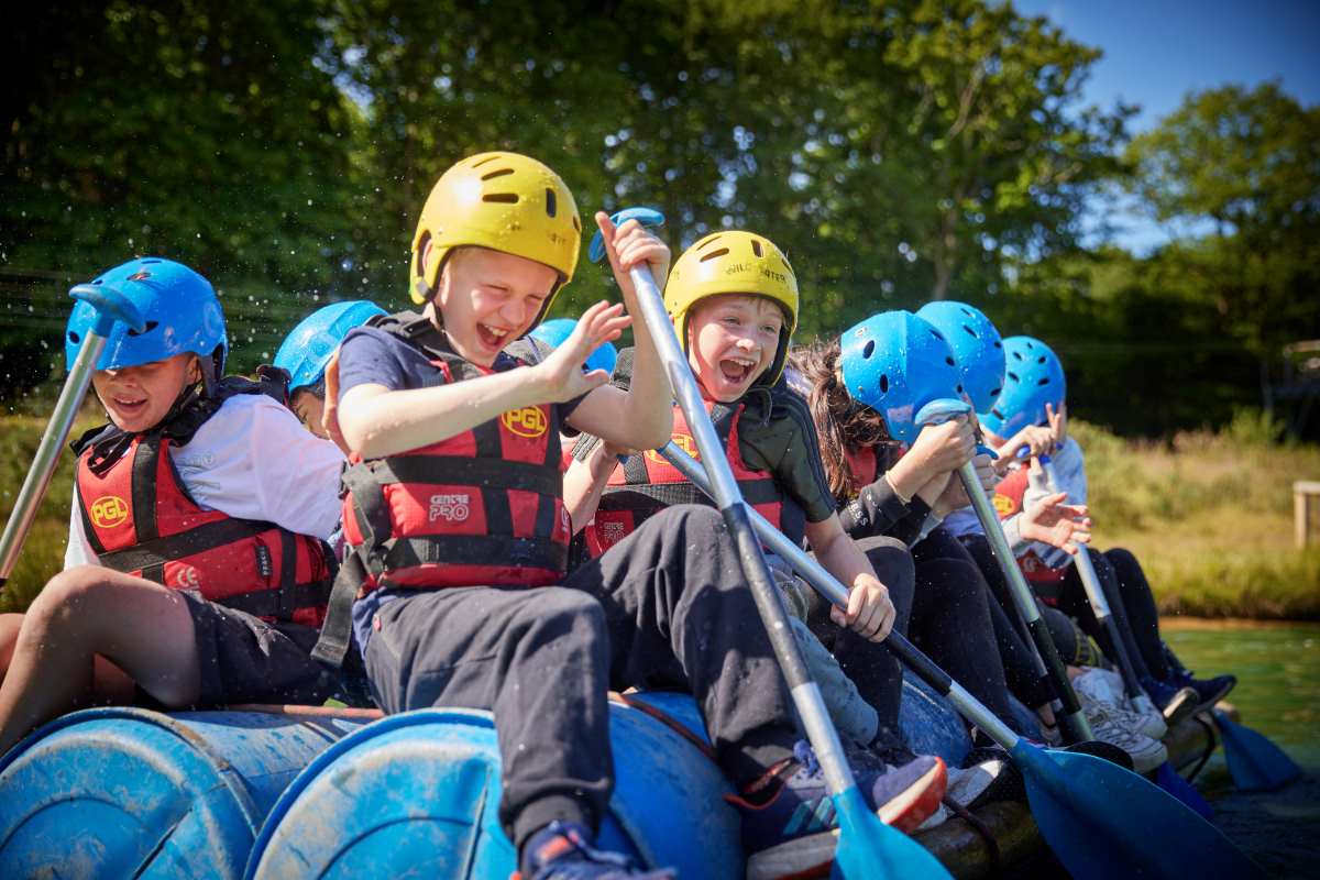 Group of children discovering the joy of rafting, wearing safety helmets and life jackets, splashing water with paddles under bright sunlight on a river.